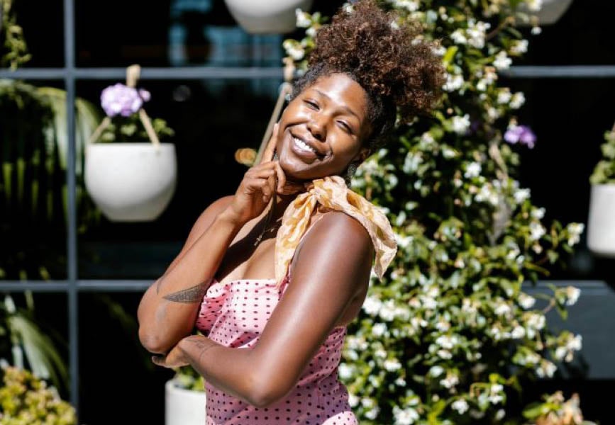 “A smiling woman stands in front of a lush vertical garden, surrounded by hanging planters and greenery.
